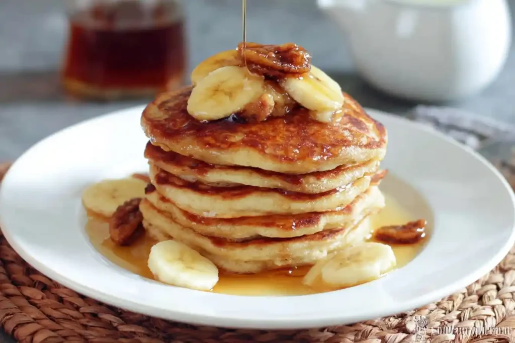 A tall stack of fluffy banana pancakes topped with sliced bananas and nuts, with syrup being drizzled from above onto a white plate.